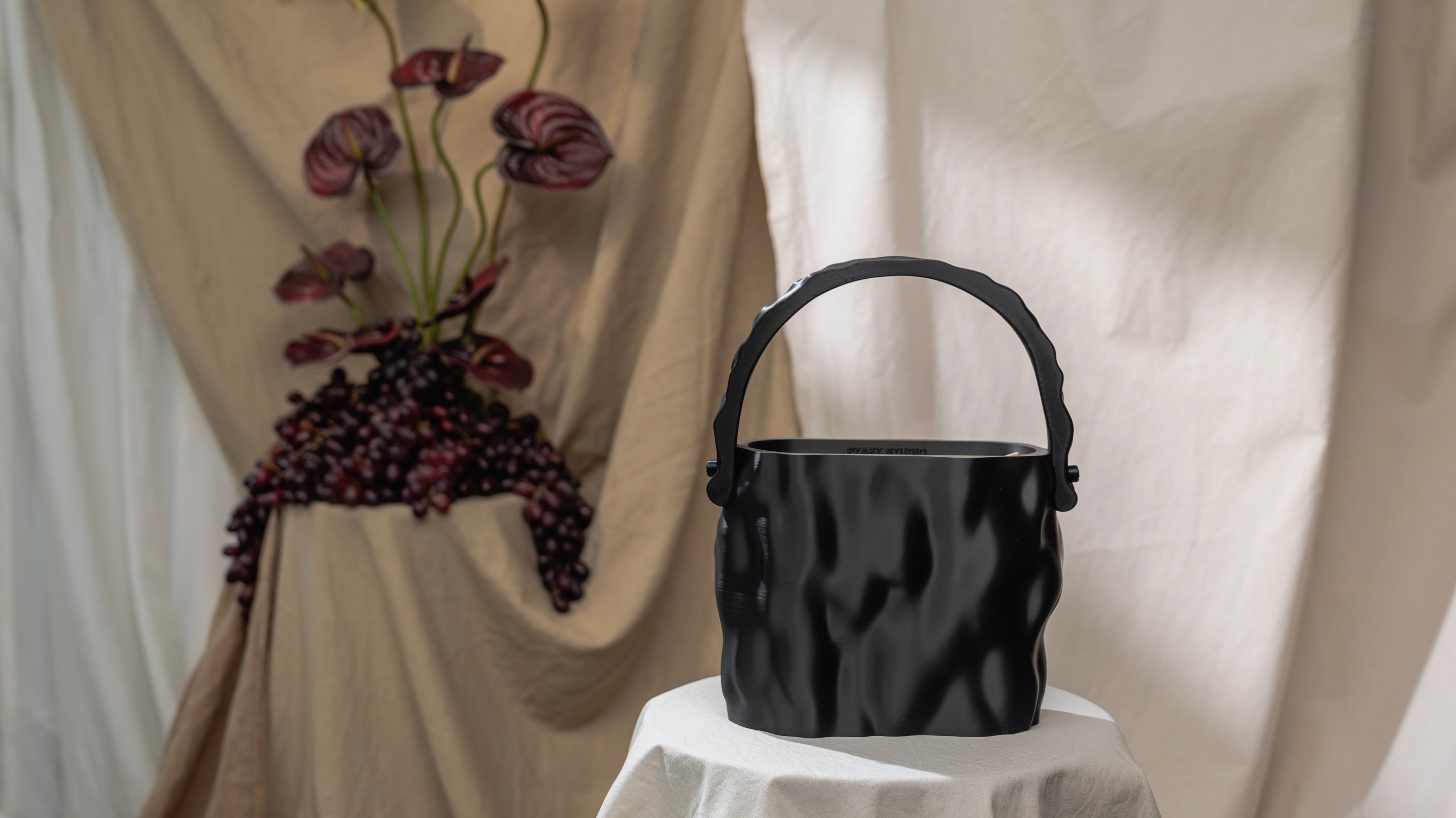 MELT bag in Obsidian placed on a draped studio set with dark grapes, sculptural flowers and soft beige fabric.