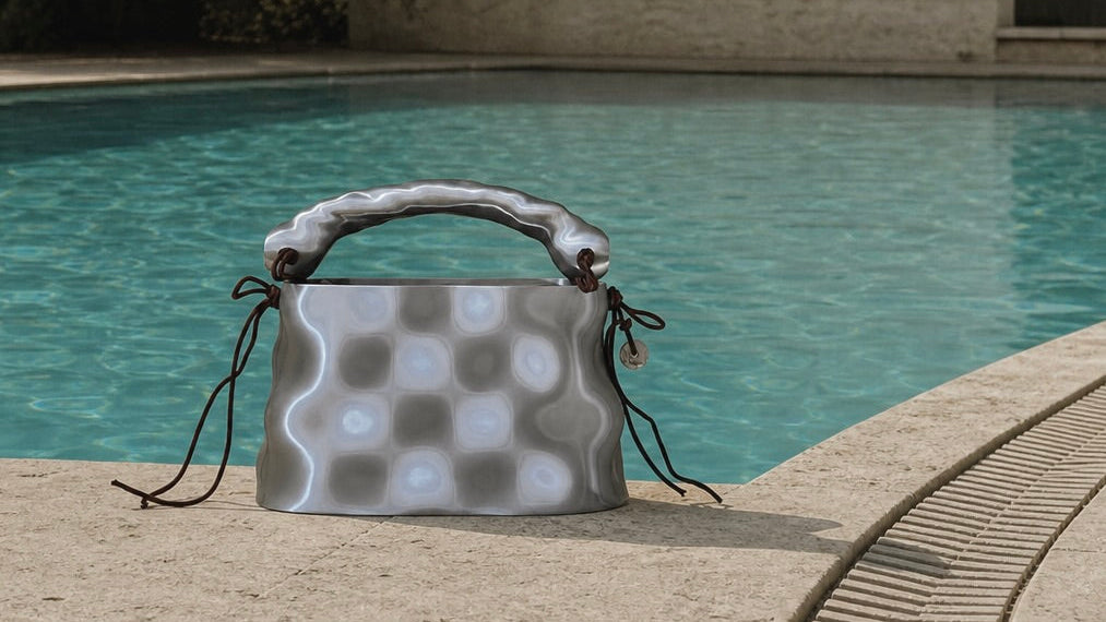 MONUMENT bag in Molten Silver placed by a pool edge with turquoise water and pale stone surroundings.