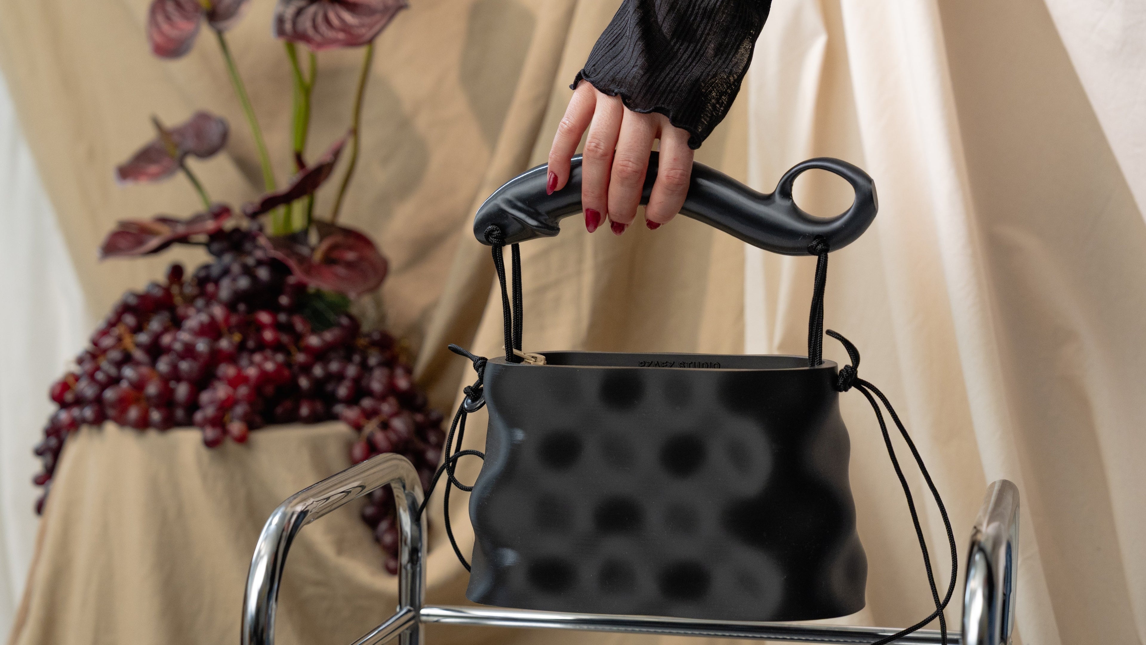 PZAEZ Softcore Obsidian black sculptural handbag held by hand, with cord details, red nails and draped fabric backdrop.