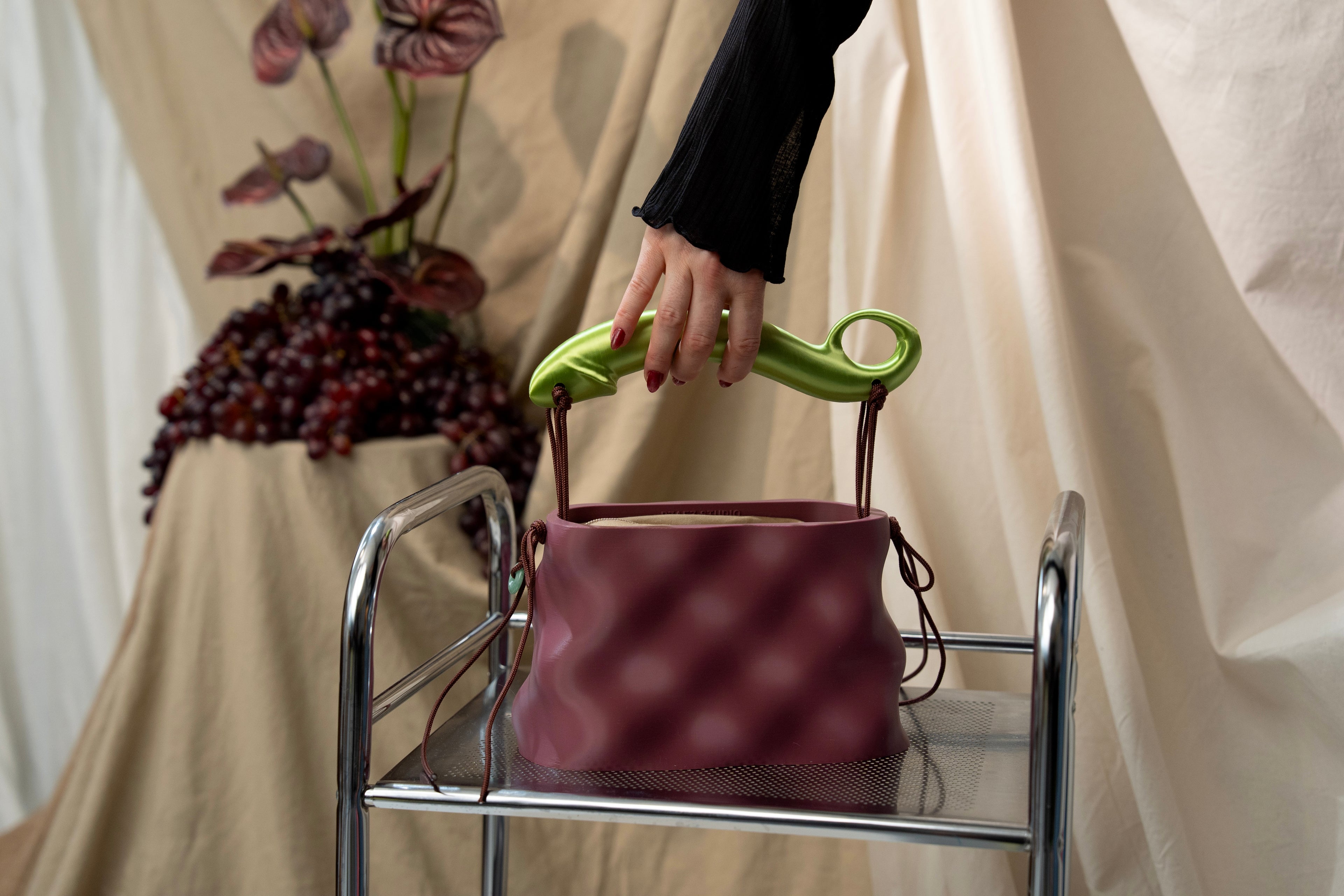 Softcore bag in Plum with green sculptural handle, held by hand on a chrome chair against draped beige fabric and grapes.
